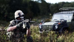 Soldat i camouflagedress knæler i højs græs med automatvåben i græ