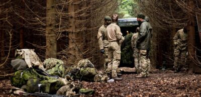 En flok soldater i camouflagedress i tæt, dansk nåleskov bag ved jeep